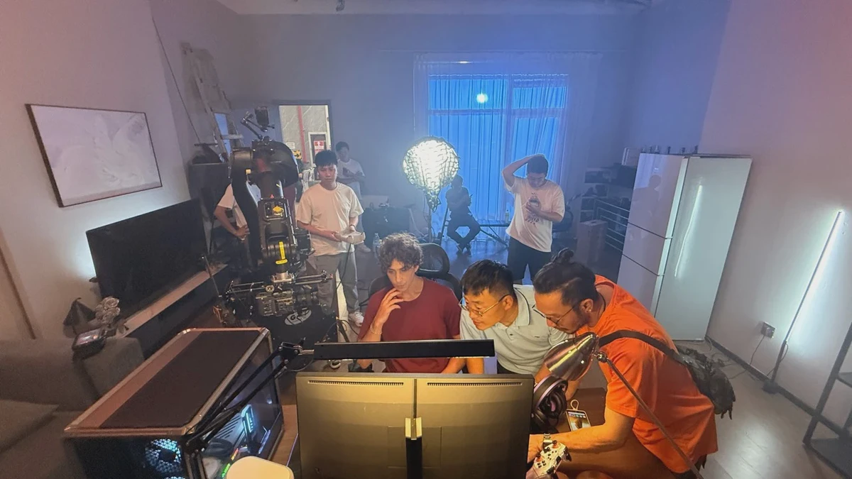 人体工学椅跨境电商视频拍摄现场，人物坐在桌前，团队在显示器旁查看画面，机械臂与灯光位于侧后方